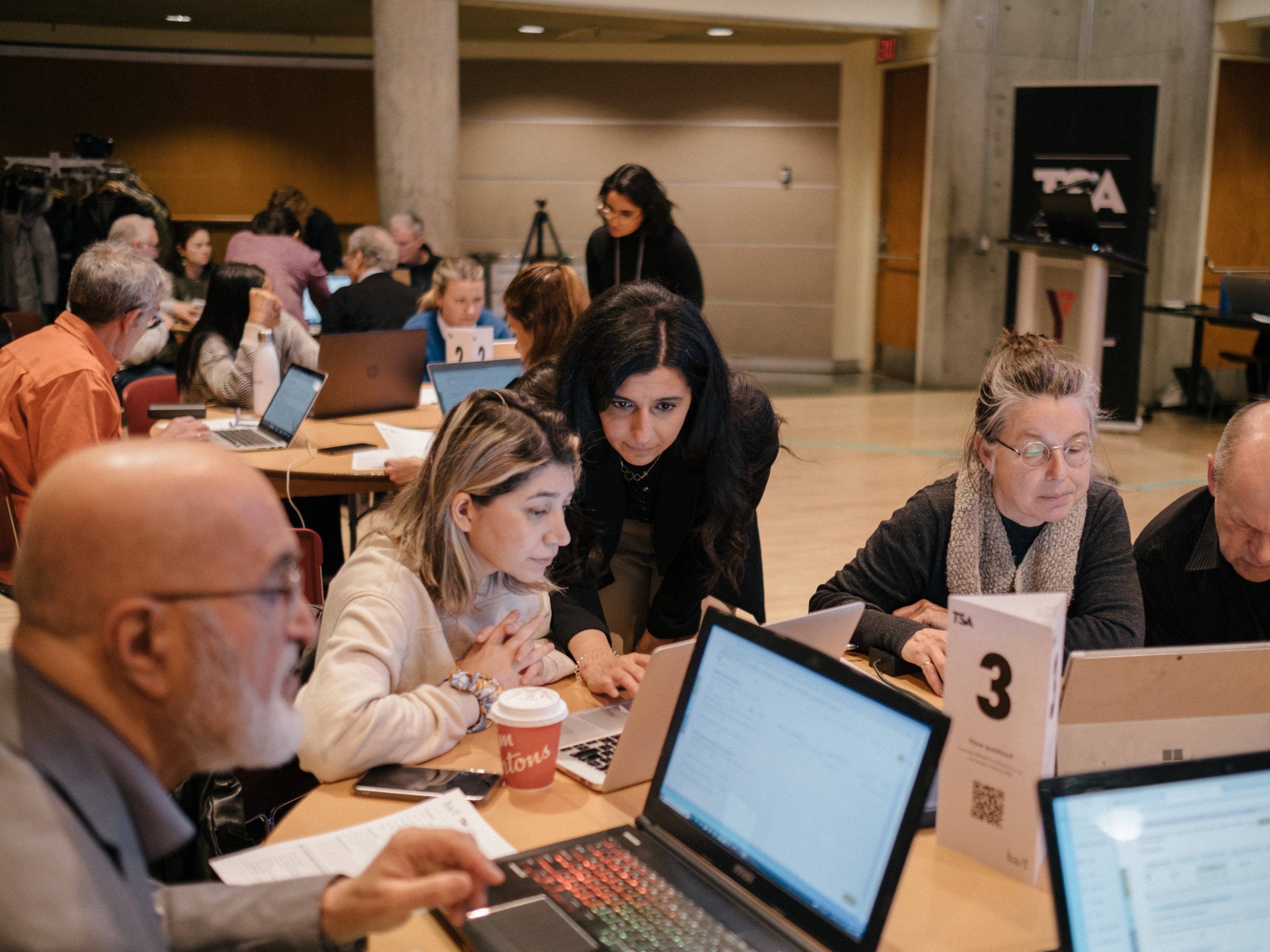 Workshop Facilitator crouches down to answer a question for the table of attendees looking intently at their laptops.