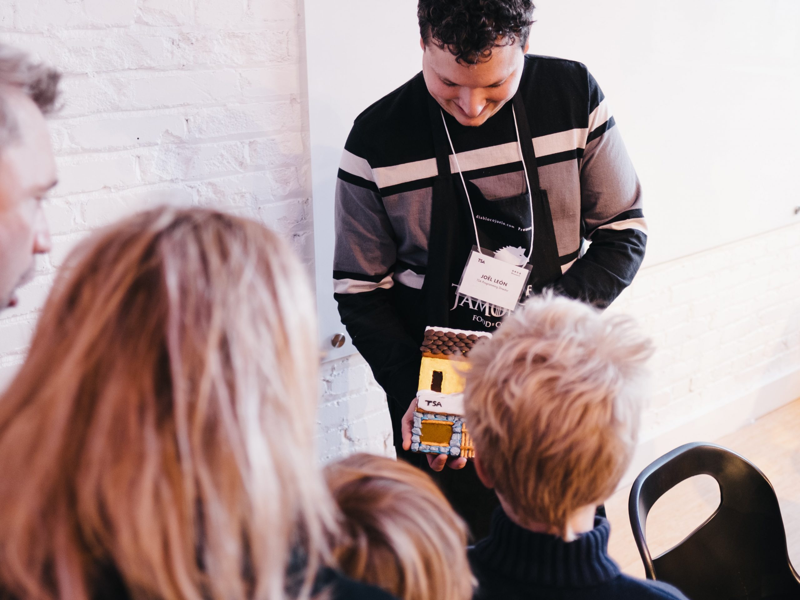 TSA Programming Director, Joël, dressed in an apron, shows off the finished example of a decorated gingerbread facade to a family with two children.